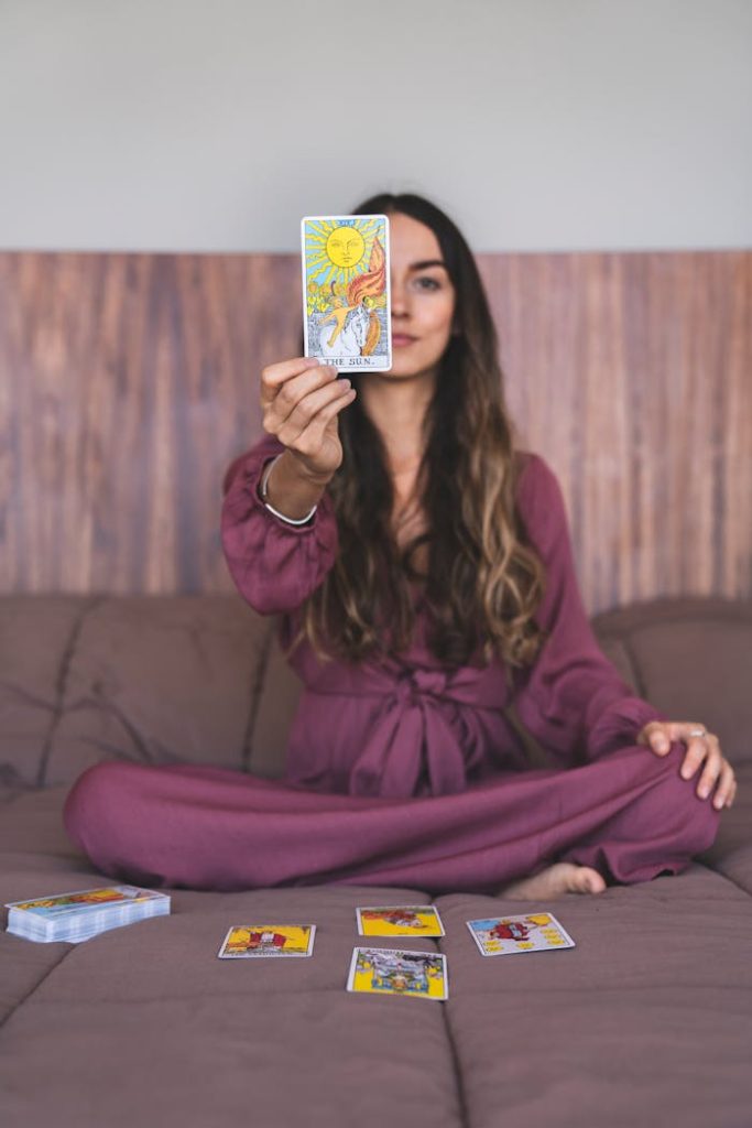 A woman sitting indoors in a purple dress holding up a tarot card, suggesting a spiritual reading.